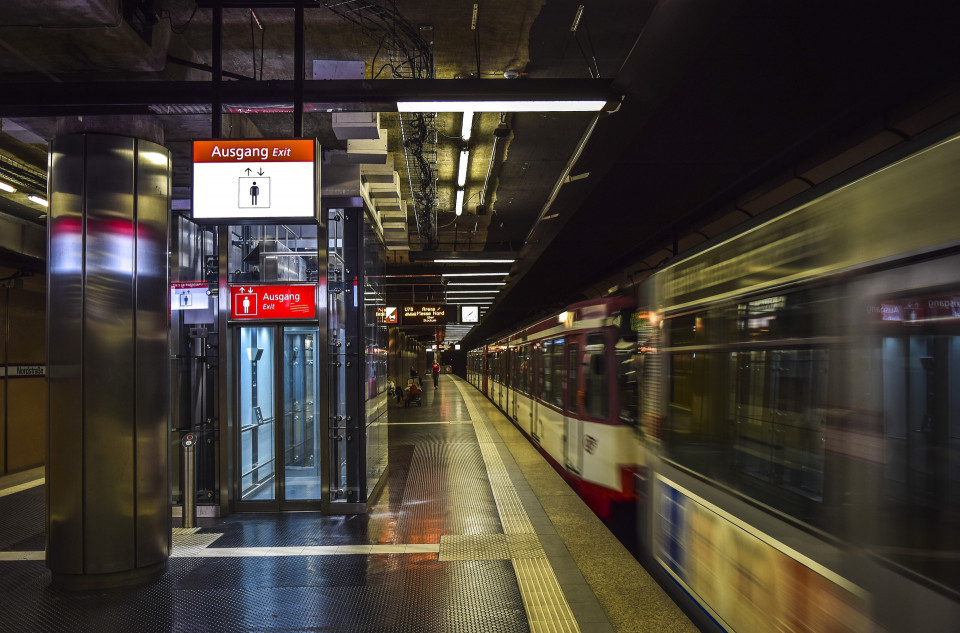 Eine unterirdische Straßenbahn-Haltestelle mit einem ausfahrenden Zug. Auf dem Bahnsteig ist ein beleuchteter Aufzug zu sehen.