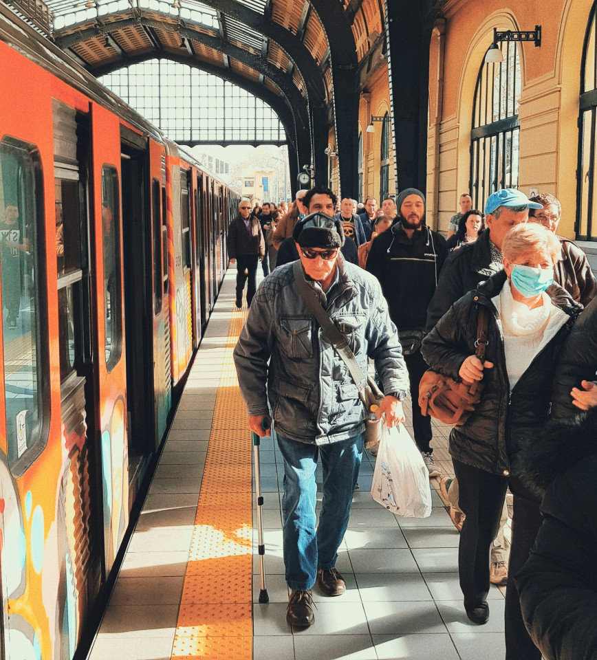 Auf einem überirdischen Seitenbahnsteig steht links ein Zug mit geöffneten Türen und rechts kommen mehrere Fahrgäste den Fotografen entgegen.