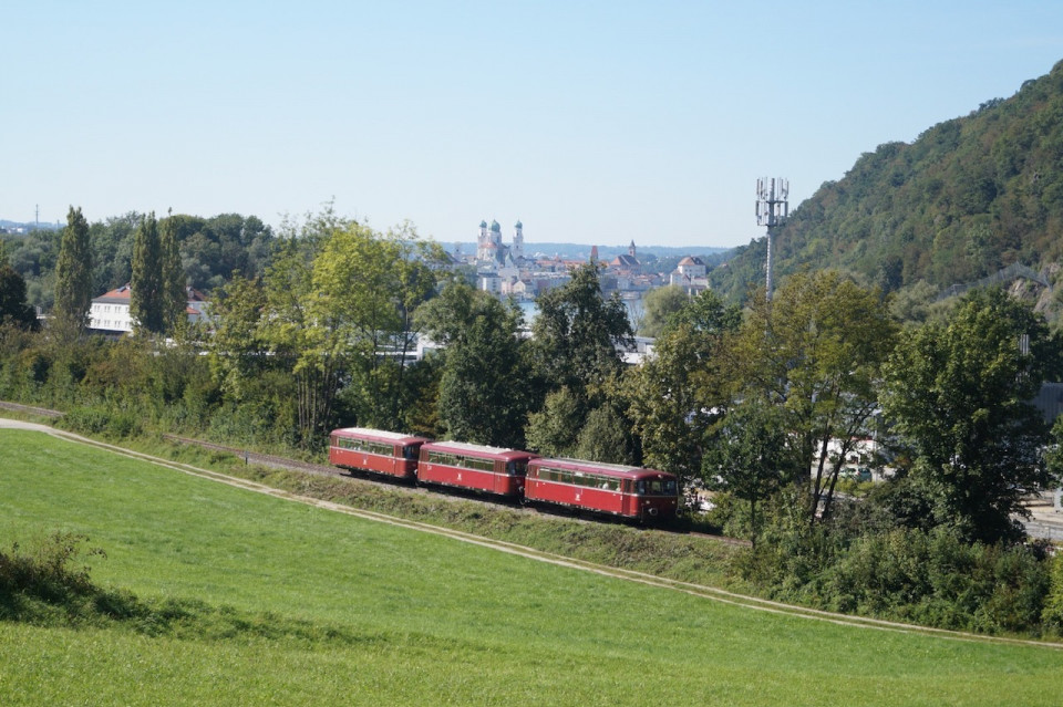 Dreiteiliger Schienenbus vor einer grünen Wiese. Im Hintergrund ist die Stadt Passau mit dem Kirchturm erkennbar. Rechts ist angeschnitten ein Berg zu sehen.