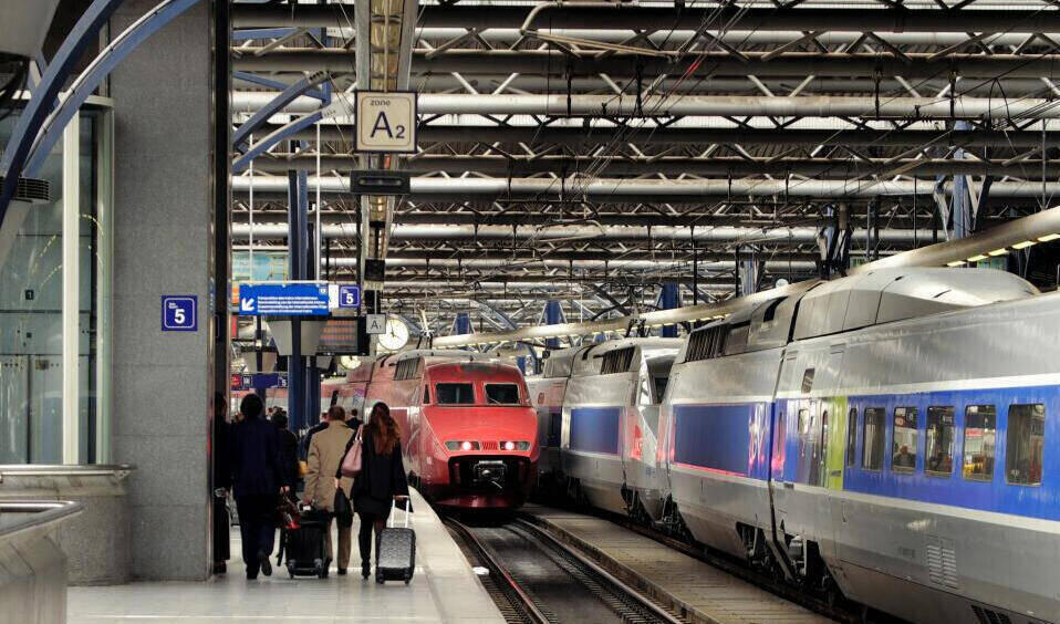 Blick auf einen Bahnsteig mit Reisenden und zwei einfahrenden Hochgeschwindigkeitszügen in einem Bahnhof mit Stahlträgerdach