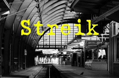 Ein Gleis in ein leeren Bahnhofshalle am Tage. Quer über das Bild steht das Wort "Streik" in gelben Buchstaben