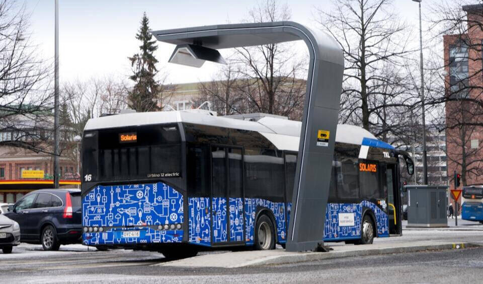 Elektrobus mit blauer, mit technischen Symbolen bedruckter Seitenfläche an einer Haltestelle unter Oberleitung in einer städtischen Umgebung mit kahlen Bäumen und mehrstöckigen Gebäuden