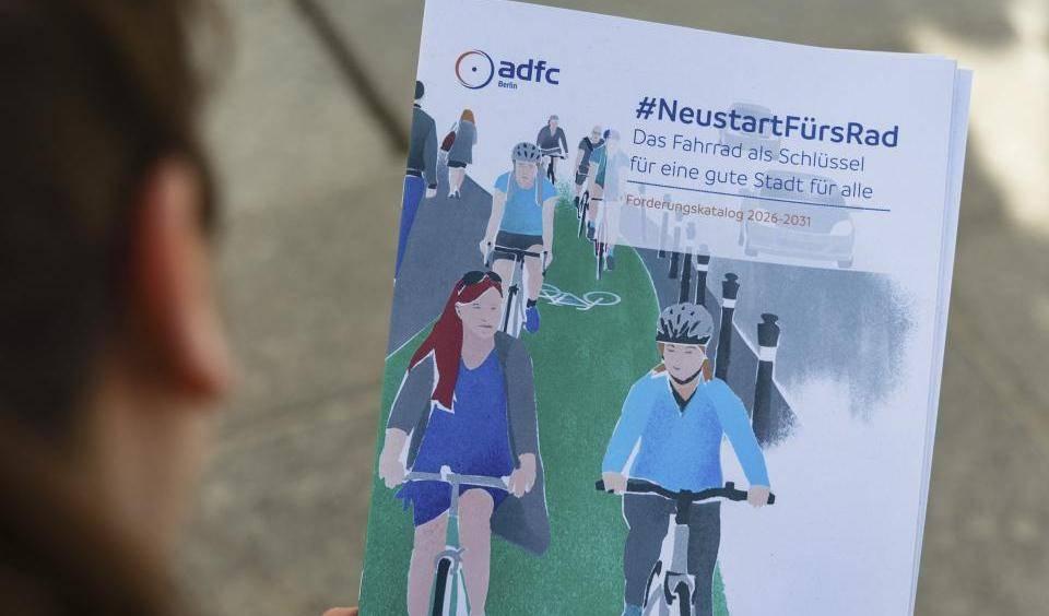 Person hält Broschüre mit Illustration von Radfahrern auf einem grünen Radweg und Text 'adfc Berlin #NeustartFürsRad Das Fahrrad als Schlüssel für eine gute Stadt für alle Forderungskatalog 2026-2031'