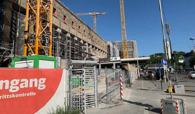 Man sieht eine große Baustelle mit mehreren Kränen und Gerüsten. Im Vordergrund befindet sich ein rotes Banner mit den Aufschriften „Eingang“ und „Zutrittskontrolle“ sowie ein Drehkreuz.