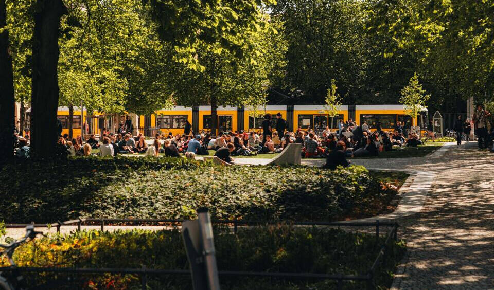 Viele Menschen sitzen auf einer Wiese unter Bäumen vor einer gelben Straßenbahn in einem Park
