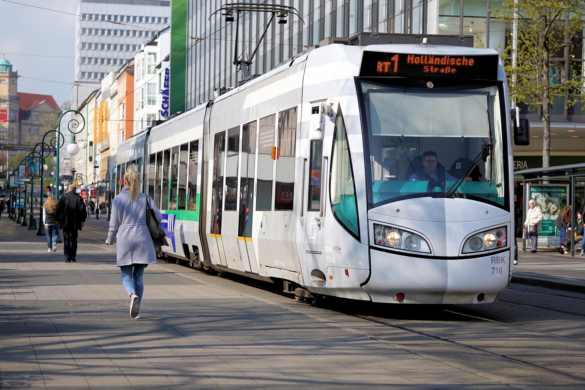 Moderner weißer Regiotram-Zug mit der Anzeige 'RT 1 Holländische Straße' fährt auf einer Stadtstraße mit Fußgängern und Gebäuden im Hintergrund. Foto: Andreas Lischka, pixabay