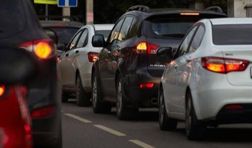 Ein Foto zeigt eine Reihe von Autos in einem Verkehrsstau, der auf einer Straße vor sich geht, mit eingeschalteten Rücklichtern und Verkehrsschildern am Rande.